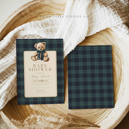 Baby shower invitation with teddy bear design on plaid patterned paper, placed on a wooden surface.