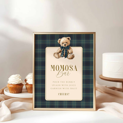 Framed sign with teddy bear illustration and text on a plaid background, placed on a surface with cupcakes and a cake.