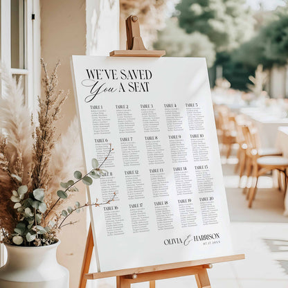 A wedding reception seating chart printed on a white board, displayed on a wooden easel in a decorated event space with pampas grass and blurred tables in the background.