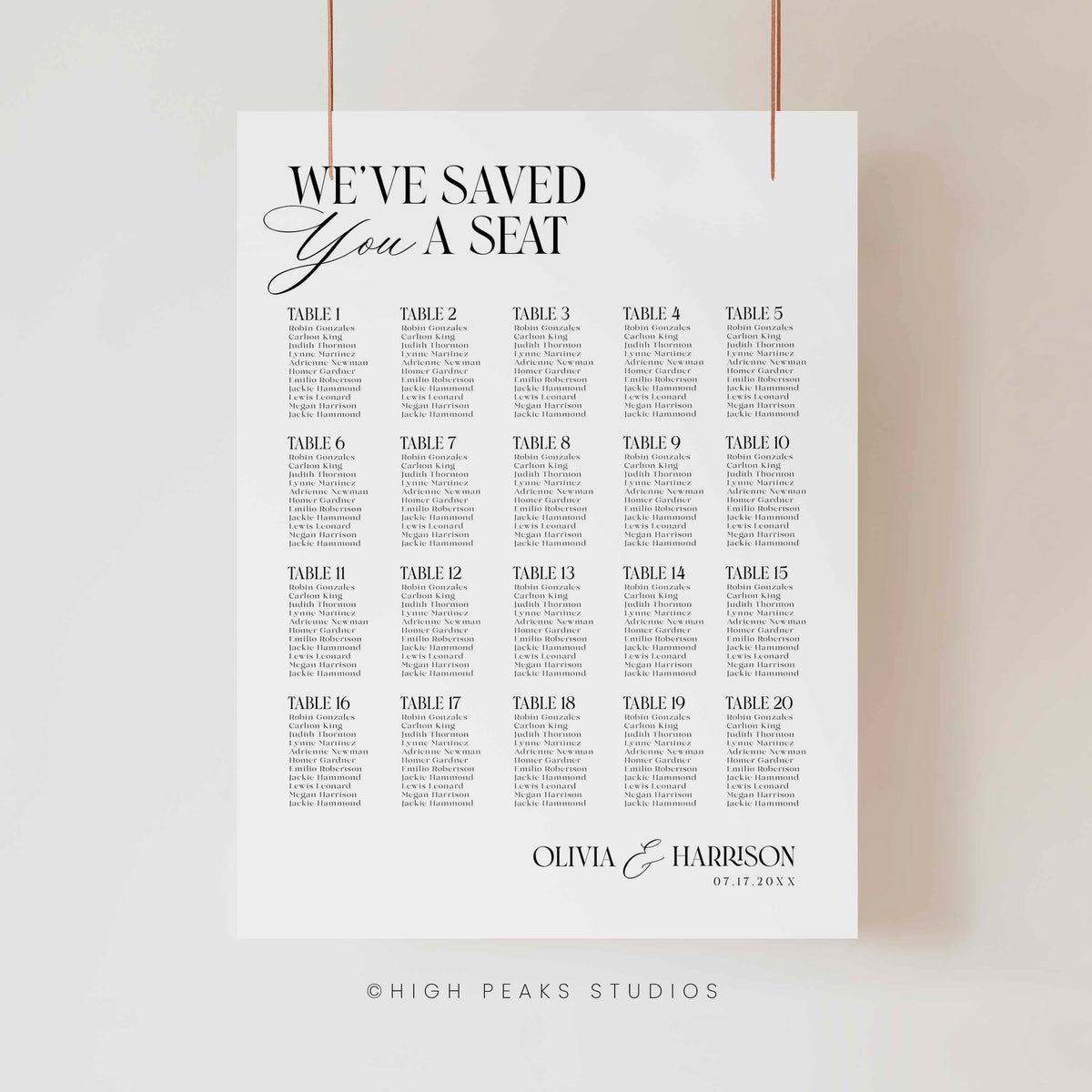 A vertical white wedding seating chart print hanging from two thin cords against a light neutral wall. The print features the text 'WE'VE SAVED YOU A SEAT' at the top, followed by a grid of table numbers from Table 1 to Table 30 with placeholder event text beneath each.