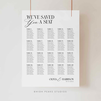 A vertical white wedding seating chart print hanging from two thin cords against a light neutral wall. The print features the text 'WE'VE SAVED YOU A SEAT' at the top, followed by a grid of table numbers from Table 1 to Table 30 with placeholder event text beneath each.
