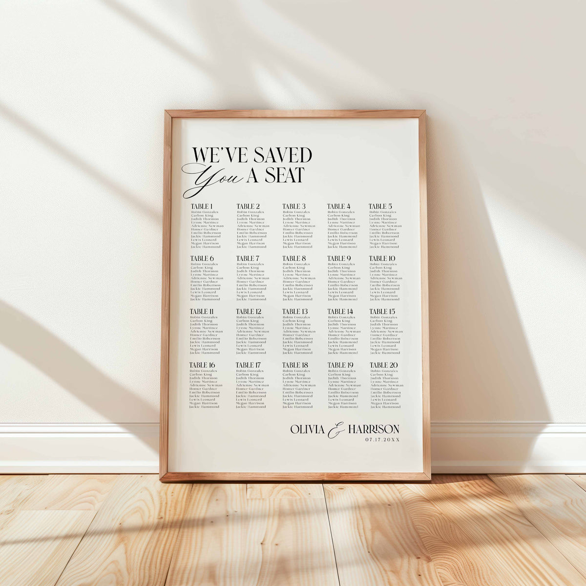 A framed wedding seating chart print leaning against a white wall on a light wood floor. The print features elegant typography reading 'WE'VE SAVED You A SEAT' followed by a grid of table assignments