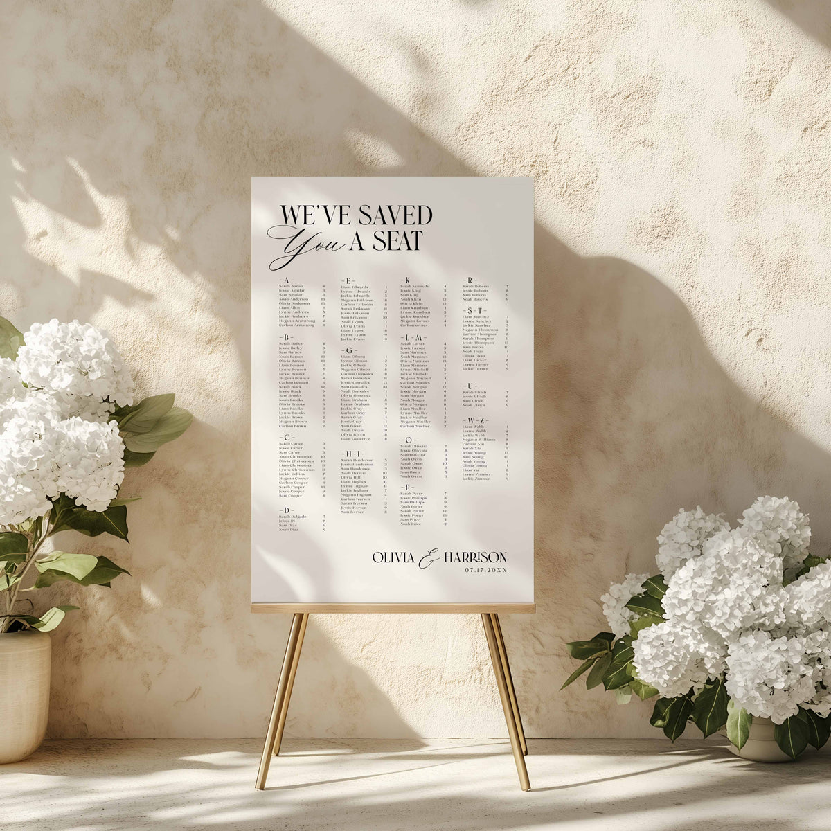 A wedding reception seating chart sign on a gold easel stand, styled on a stone floor with white flowers on either side.