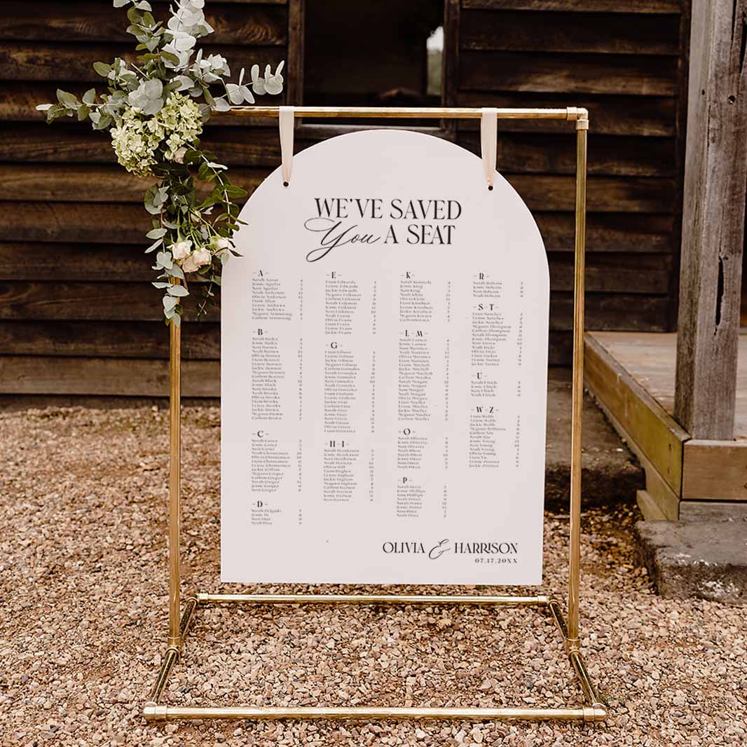 A gold metal freestanding arch table stand holding a white arch-shaped wedding seating chart with black text, photographed outdoors on gravel in front of a rustic wooden fence. A floral arrangement hangs from the left side of the stand.