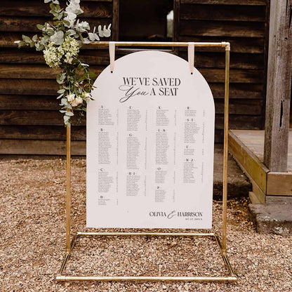 A gold metal freestanding arch table stand holding a white arch-shaped wedding seating chart with black text, photographed outdoors on gravel in front of a rustic wooden fence. A floral arrangement hangs from the left side of the stand.