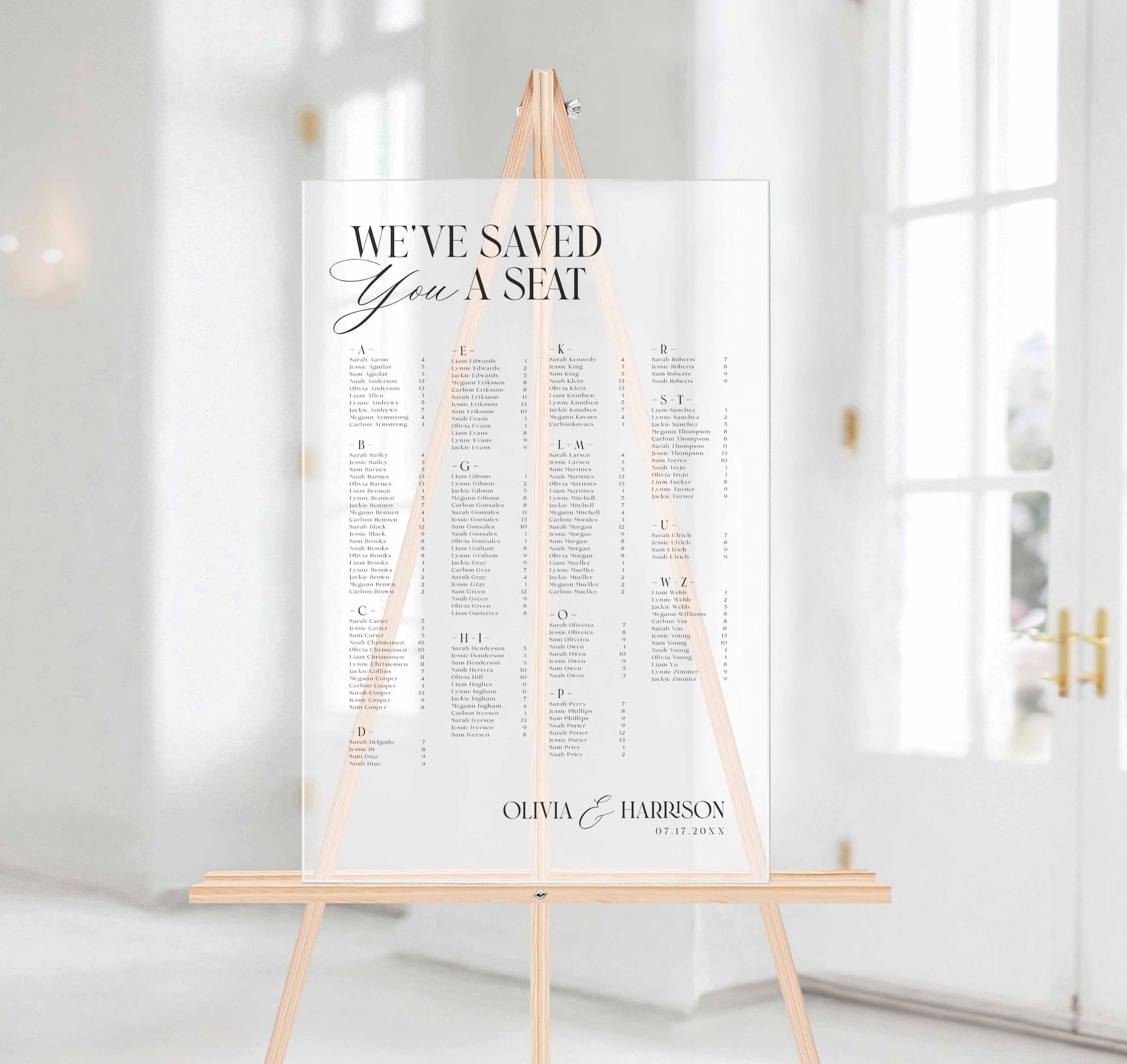 A wedding seating chart printed on a frosted acrylic board, displayed on a light wood easel in a bright, modern white hallway.