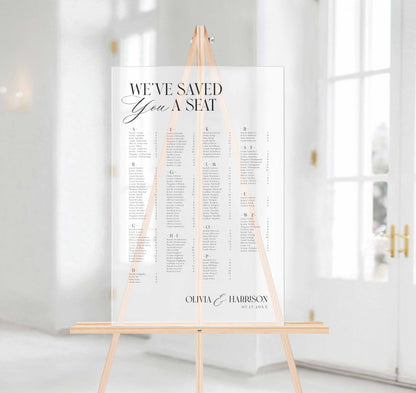 A wedding seating chart printed on a frosted acrylic board, displayed on a light wood easel in a bright, modern white hallway.