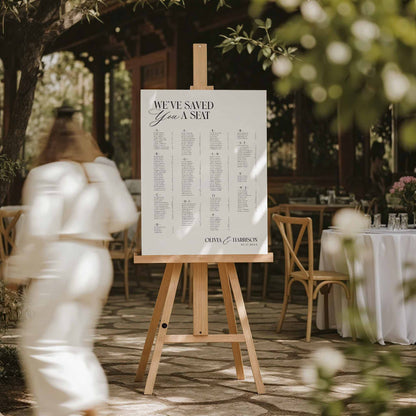 A wedding guest list sign reading 'We've Saved You a Seat' displayed on a wooden easel in an outdoor garden event setting. A blurred person in a white outfit walks past in the foreground, with tables and chairs visible in the background.