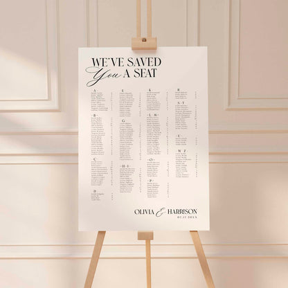 A wedding seating chart print displayed on a wooden easel in a softly lit interior with cream paneled walls. The print reads “WE’VE SAVED You A SEAT” at the top and includes a seated list for a wedding couple.
