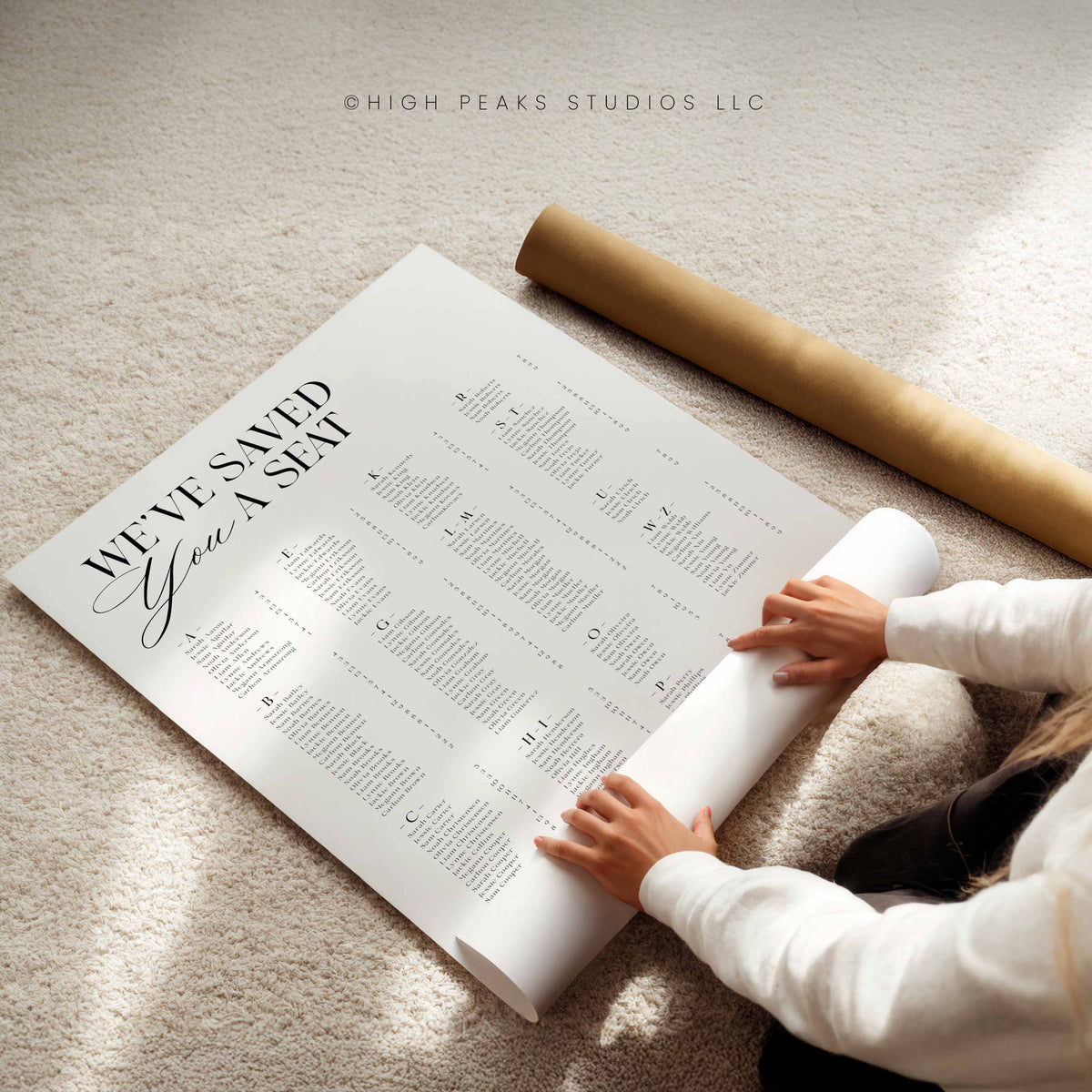A person unrolling a large printed poster on a beige carpet, with a brown cardboard mailing tube nearby. The poster reads “WE’VE SAVED YOU A SEAT” with dense black text.