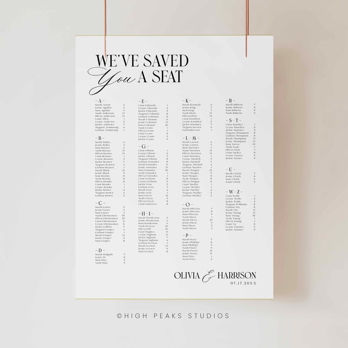 A vertical white wedding seating chart print clipped against a light neutral wall, with black typography reading 'WE'VE SAVED YOU A SEAT' and a personalized list section at the bottom. Thin copper-colored hanging cords are visible at the top, and 'HIGH PEAKS STUDIOS' appears faintly below the print.