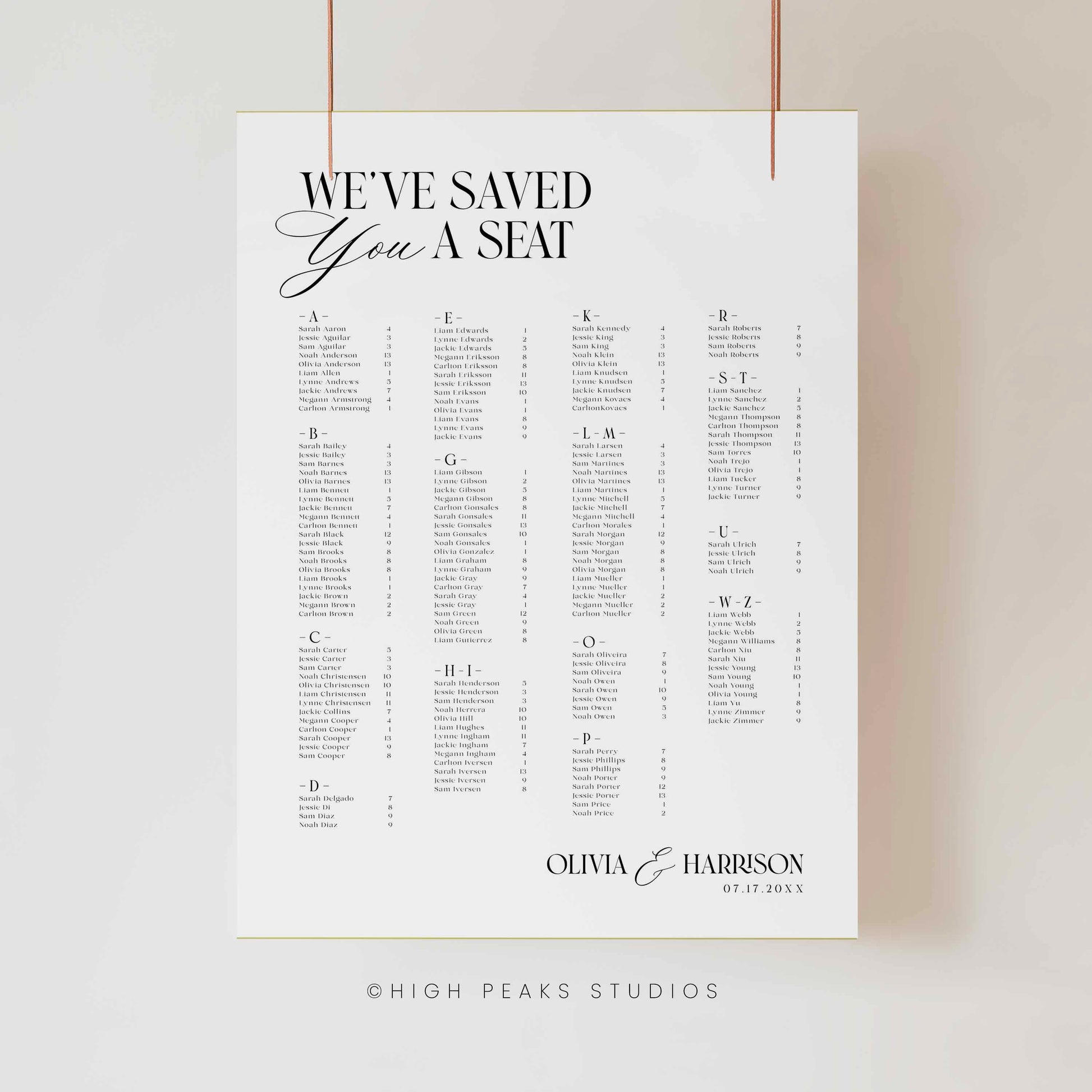 A vertical white wedding seating chart print clipped against a light neutral wall, with black typography reading 'WE'VE SAVED YOU A SEAT' and a personalized list section at the bottom. Thin copper-colored hanging cords are visible at the top, and 'HIGH PEAKS STUDIOS' appears faintly below the print.