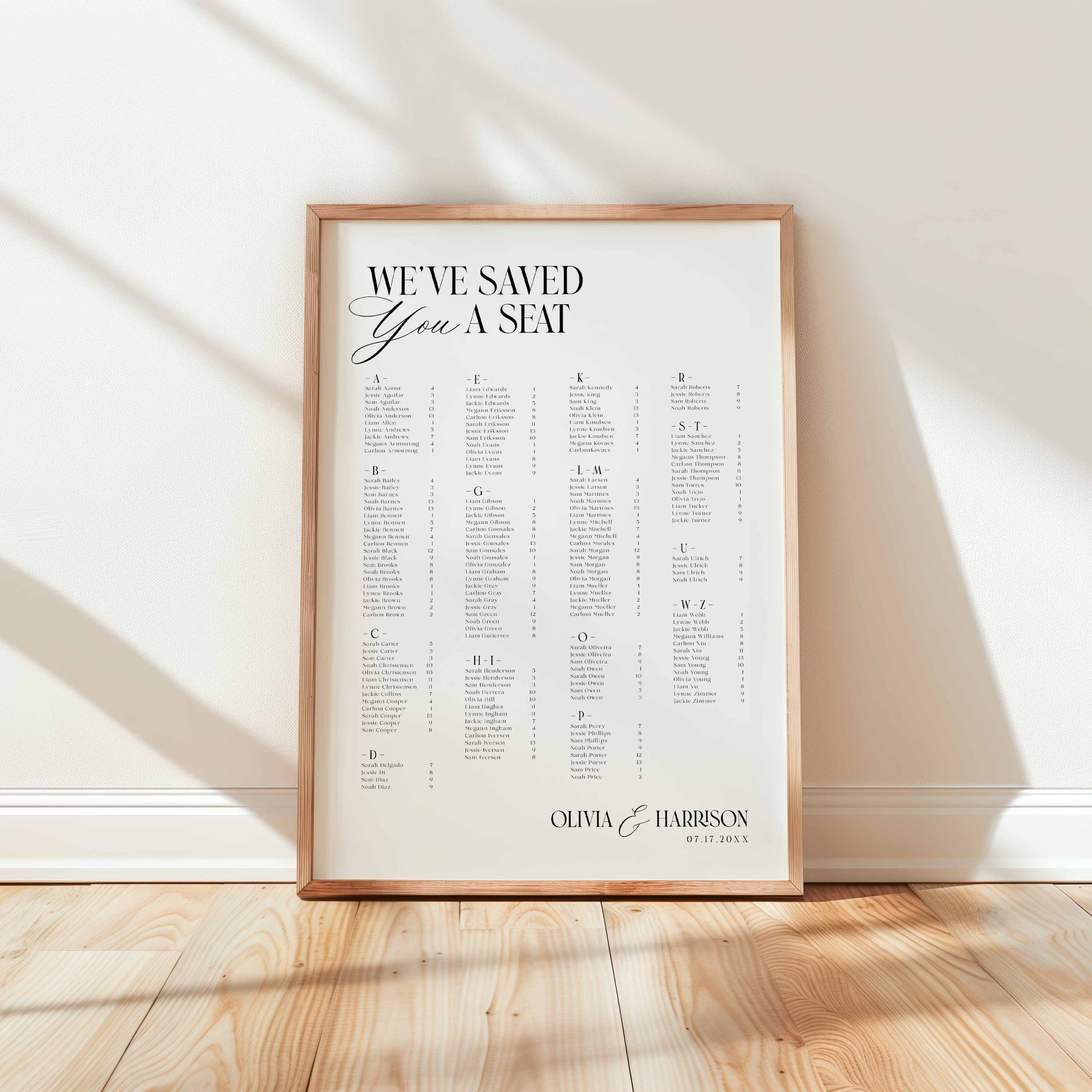 A framed personalized seating chart print leaning against a white wall on a light wood floor. The print features the text 'WE'VE SAVED YOU A SEAT' at the top, followed by a grid of alphabetized names.