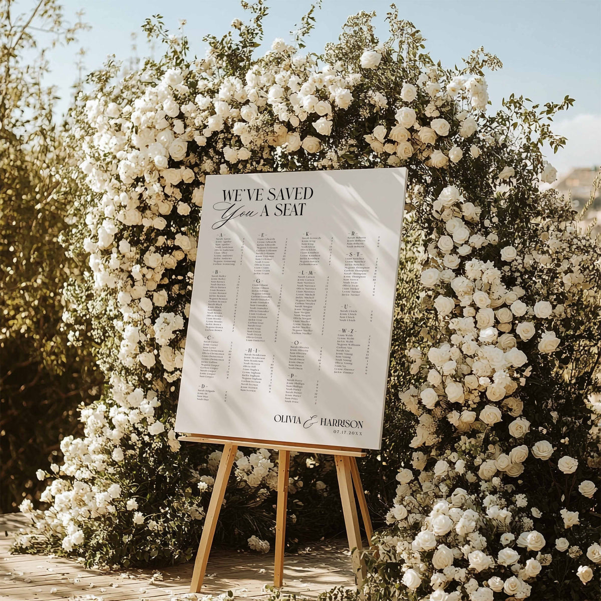 A wedding guest seating sign on a wooden easel, displayed outdoors in front of a large floral arch of white roses. The sign reads 'WE'VE SAVED Your A SEAT' and features the names' of the wedding couple
