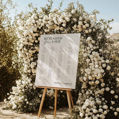 A wedding guest seating sign on a wooden easel, displayed outdoors in front of a large floral arch of white roses. The sign reads 'WE'VE SAVED Your A SEAT' and features the names' of the wedding couple