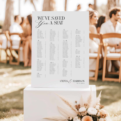 A wedding seating chart sign reading 'WE'VE SAVED YOU A SEAT' with the names Olivia and Harrison, displayed on a white pedestal in an outdoor wedding setting with guests seated in the background.