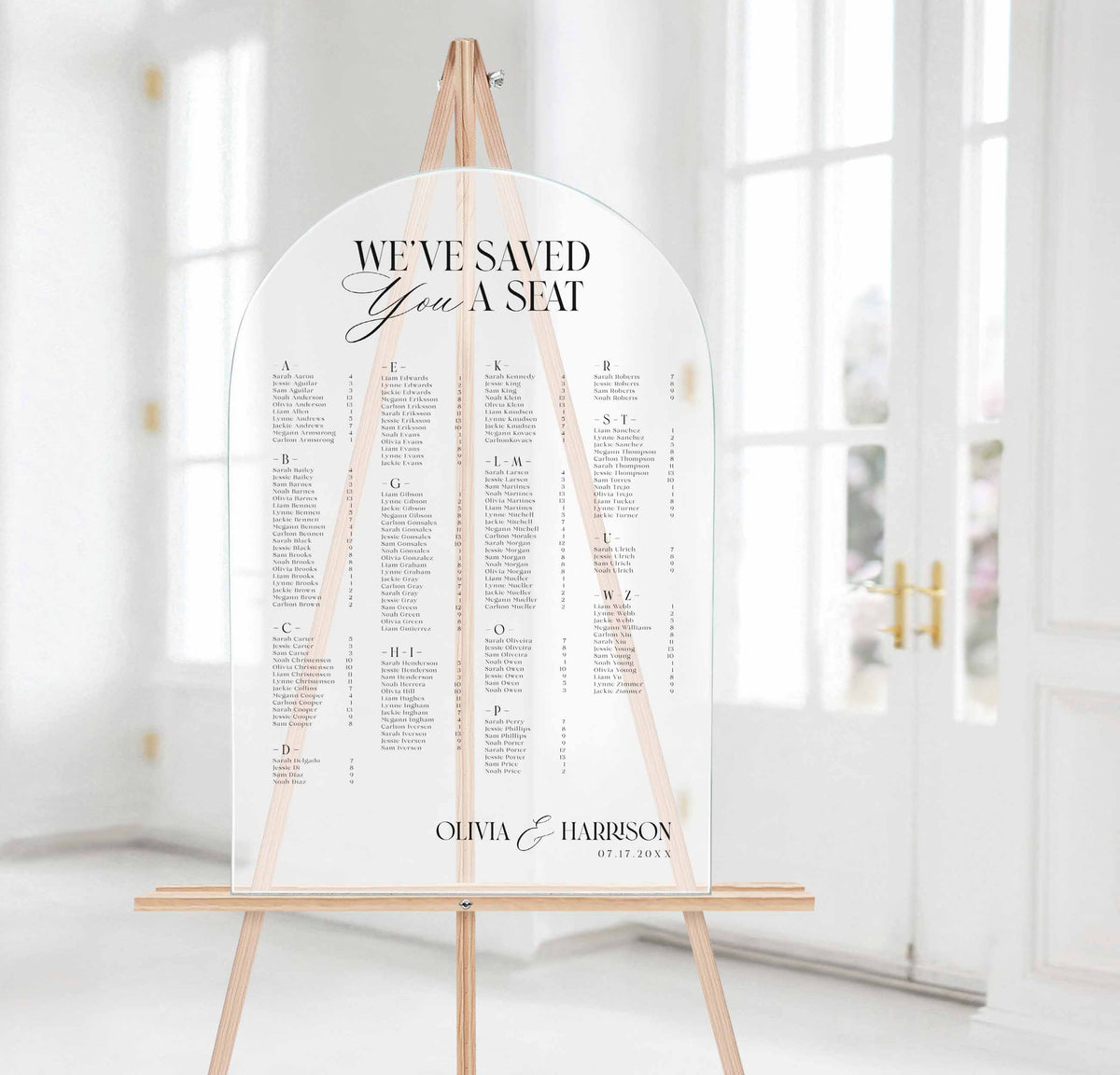 A wedding seating chart sign with an arched white backdrop and black text, displayed on a wooden easel in a bright, softly blurred indoor setting with white doors in the background.